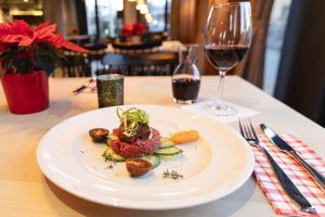 Rindertatar mit Kräutern und Gurken auf einem weißen Teller neben einem Glas Rotwein.