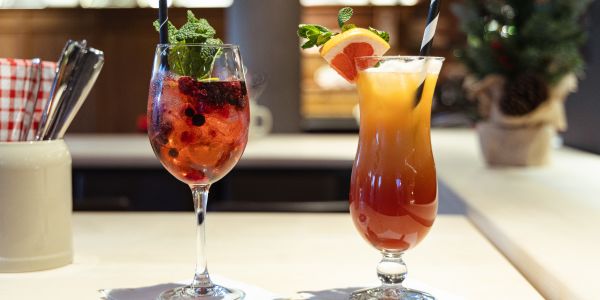 Zwei Cocktails in Gläsern, einer mit Beeren und einer mit Orange, auf einer hellen Theke.