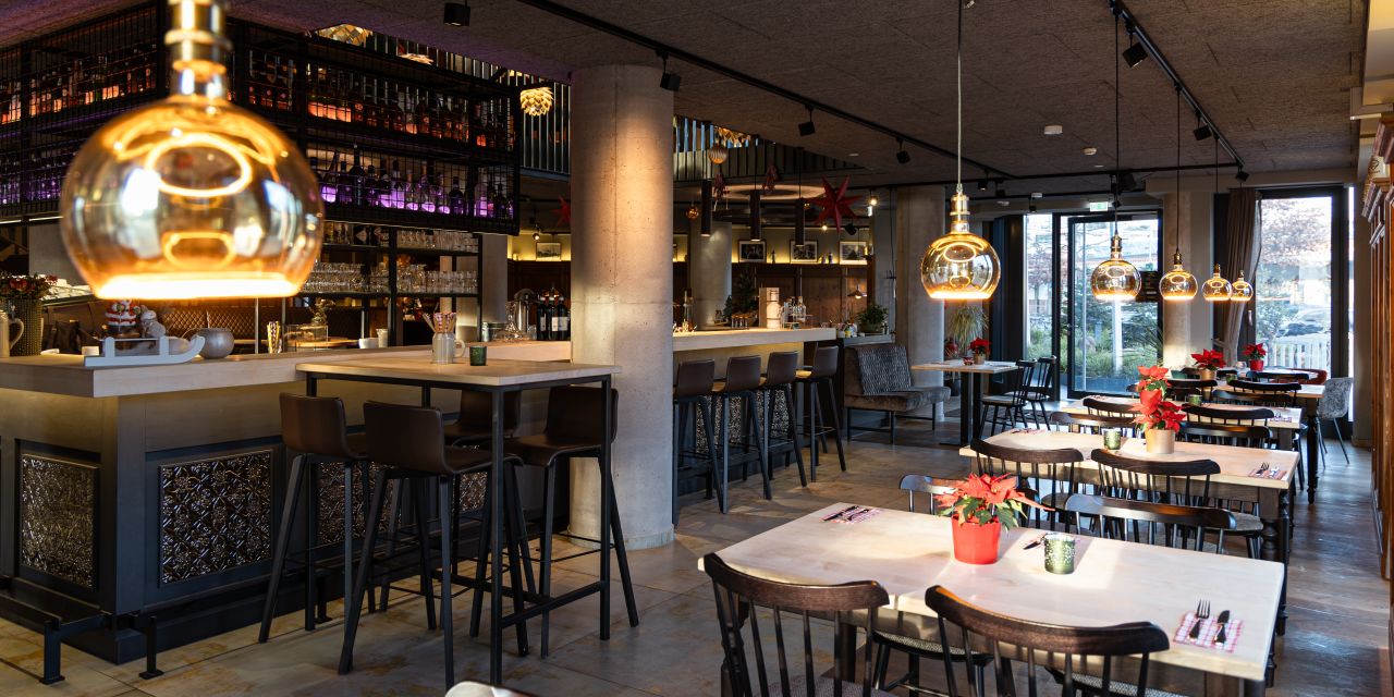 Gastraum mit modernen Pendelleuchten, Bartischen und festlich dekorierten Esstischen.